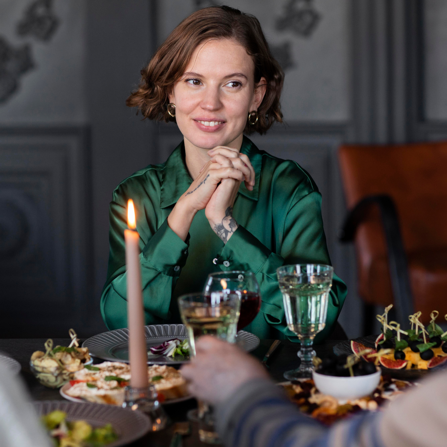 Woman in a green jacket sitting at a table with a candle and wine glasses, smiling.